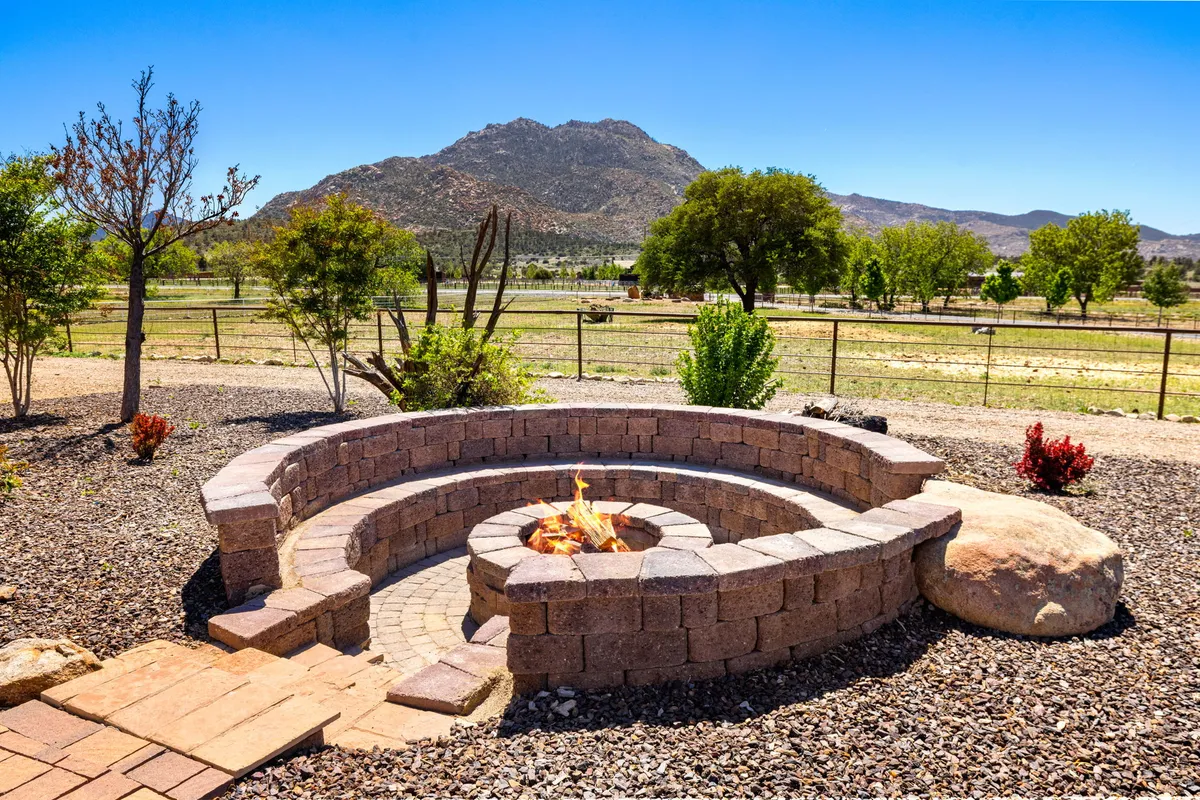 This image captures an inviting fire pit area set against a dramatic mountain backdrop, surrounded by natural vegetation and a pathway leading toward open outdoor spaces. The scene evokes tranquility, with the Arizona wilderness and rugged granite formations visible in the distance.
