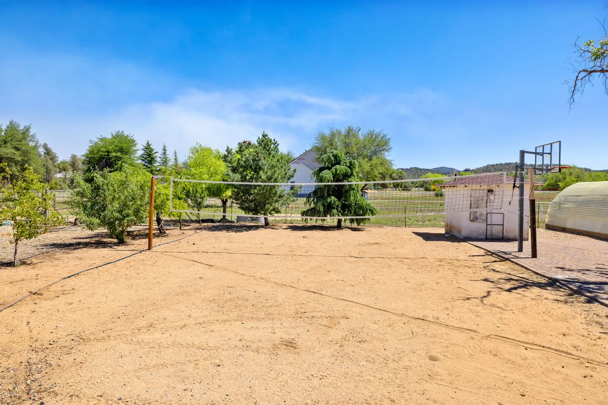 This exterior image captures a sandy open area with a volleyball net, basketball hoop, and is framed by mature trees. The backdrop is expansive and natural, providing a sense of outdoor space ideal for gathering, playing, and socializing. The rustic setting hints at the ranch environment and possibility for al fresco activities.