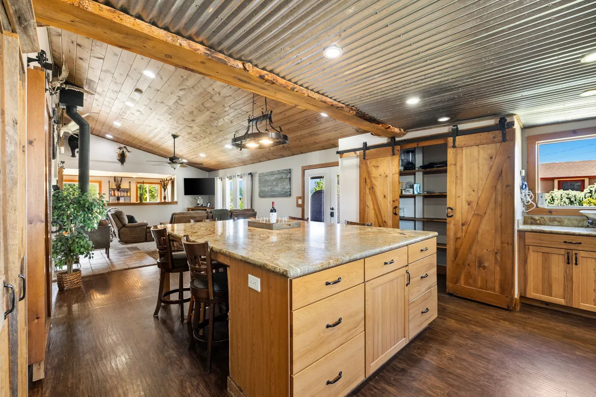 This image showcases a spacious open-plan kitchen with a large kitchen island, rustic wooden cabinetry, an open living area visible in the background, and inviting light fixtures overhead. The atmosphere is warm and inviting, suggesting a space where family and friends can gather for cooking, conversation, and shared meals.