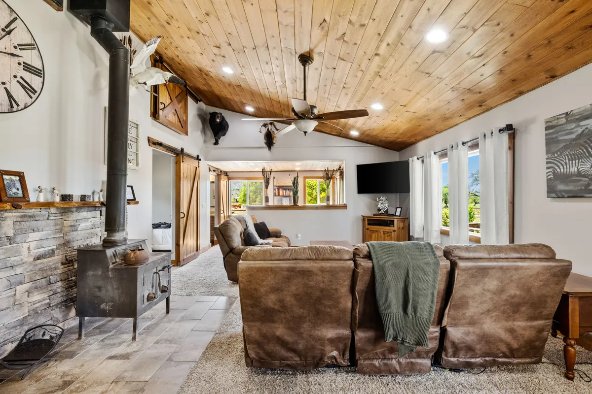 A warm, welcoming living area with leather sofas, a striking stone fireplace, wooden ceiling, and expansive windows letting in abundant natural light and offering views of the ranch. The arrangement encourages both relaxation and togetherness while maintaining a strong connection to the outdoors.