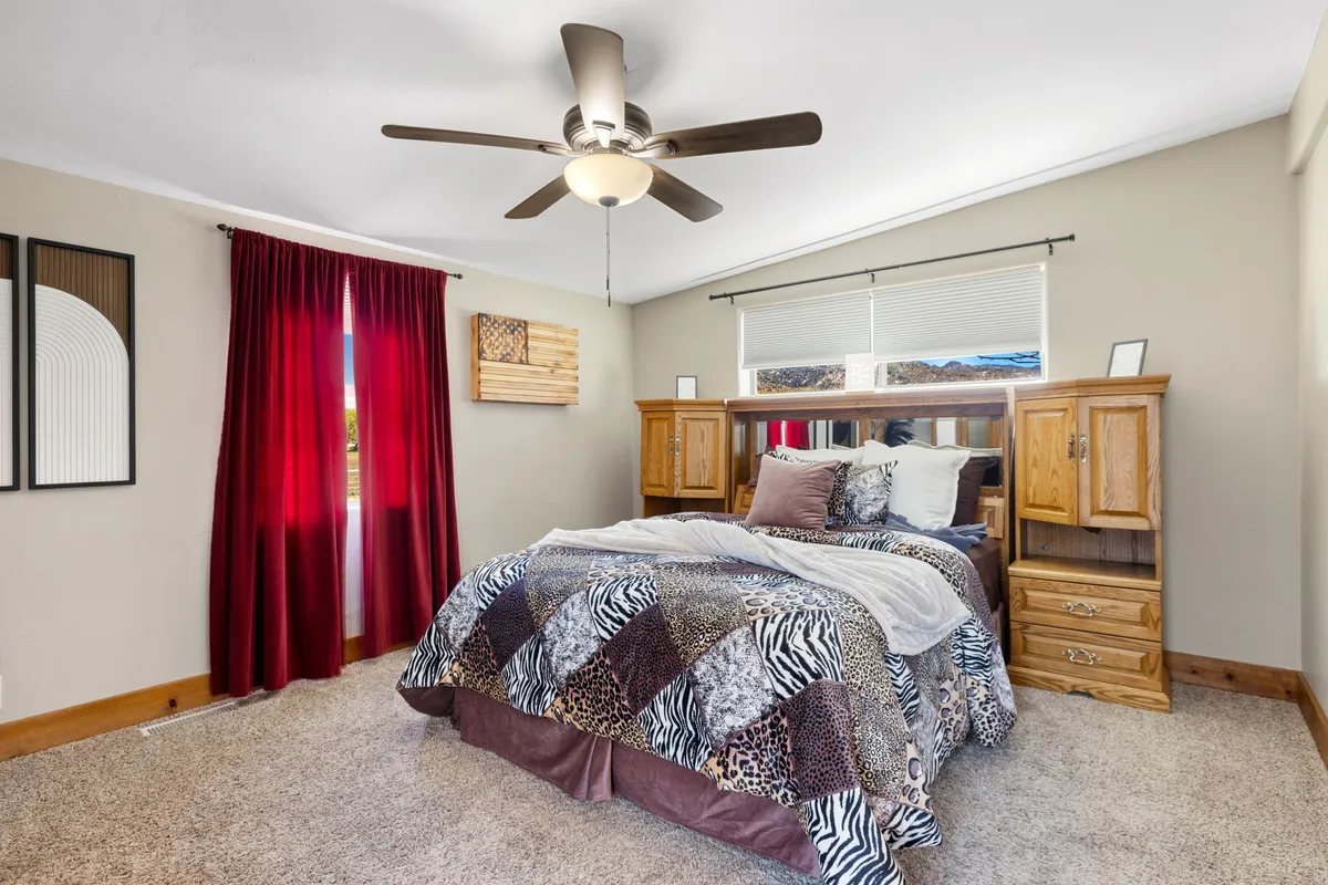 A warmly decorated bedroom with patterned bedding, red curtains, wooden furniture, and wall art. The bed is positioned to catch the morning sunlight, creating an inviting setting for rest and relaxation.
