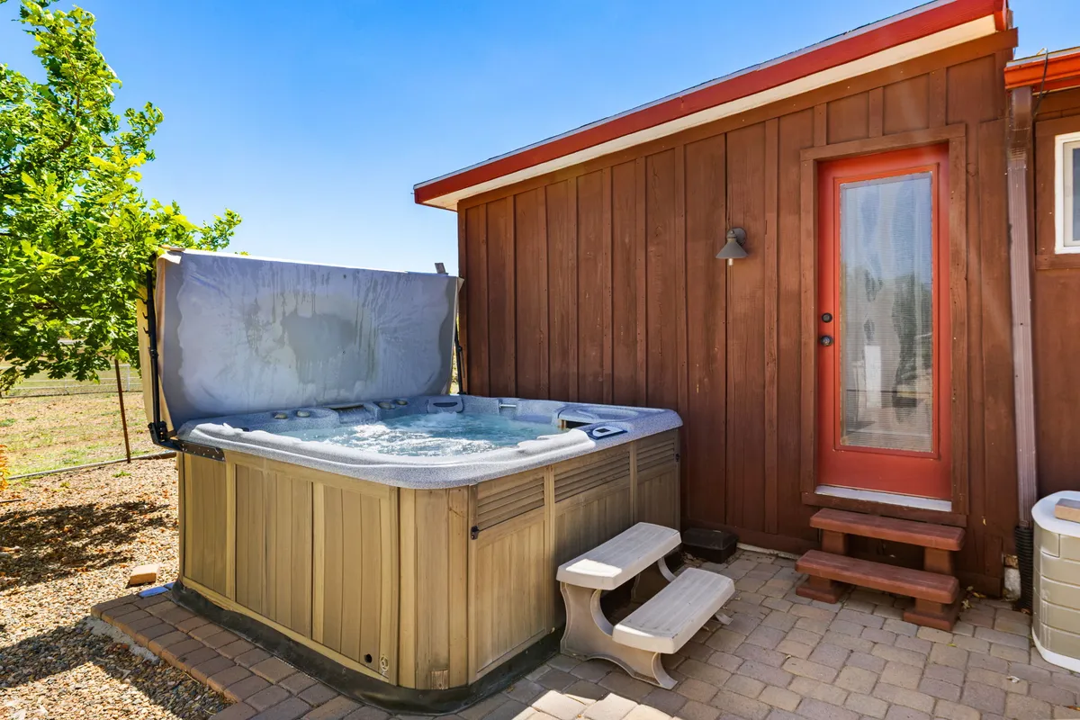This image showcases a bubbling hot tub situated on a stone patio adjacent to the wooden siding of the main house, with a red door adding a pop of color. The patio offers relaxing seating and is surrounded by natural elements, hinting at open views beyond.