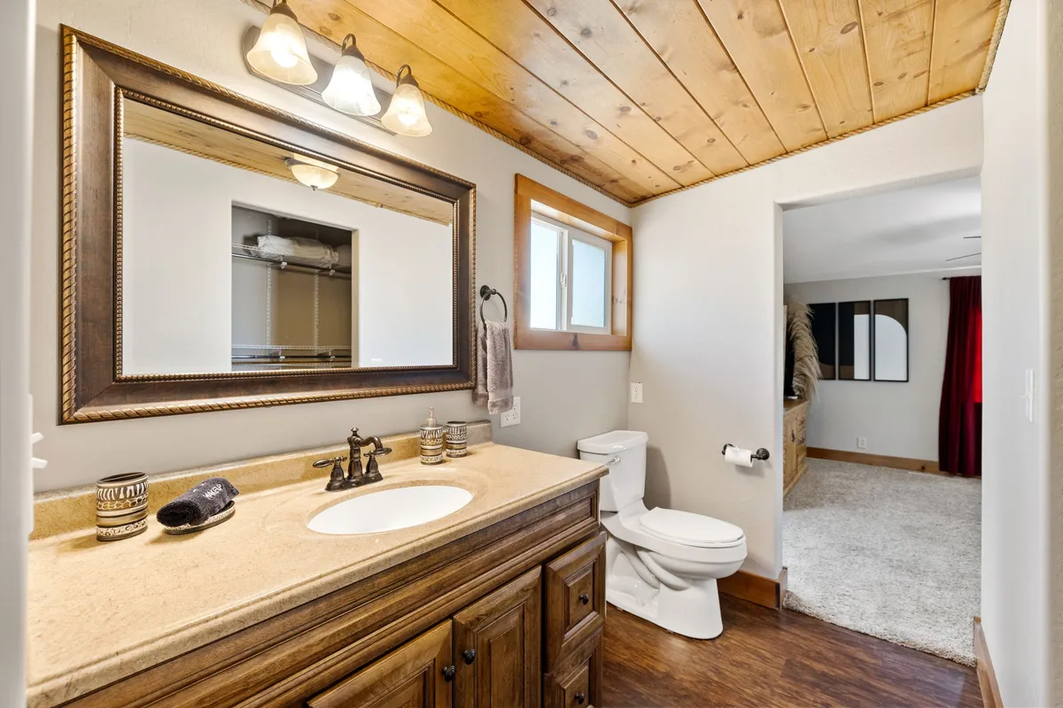 A modern bathroom featuring a vanity with a sink, large mirror, wooden ceiling, and a window. The space is bathed in natural light, making it functional yet soothing for daily routines.