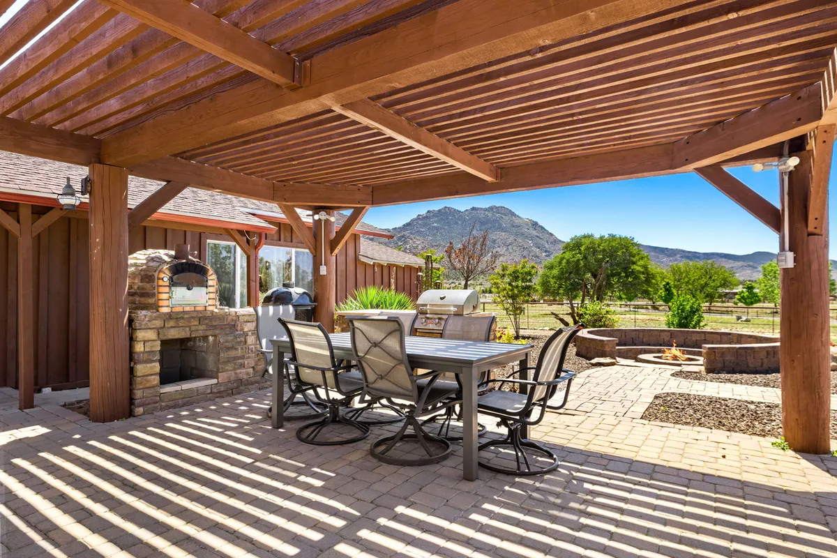 This exterior photo features an outdoor dining area with a table and chairs set on a patio, accompanied by an outdoor kitchen grill, a stone pizza oven, and a fire pit. The backdrop showcases a beautiful mountain view, enhancing the feeling of outdoor living.