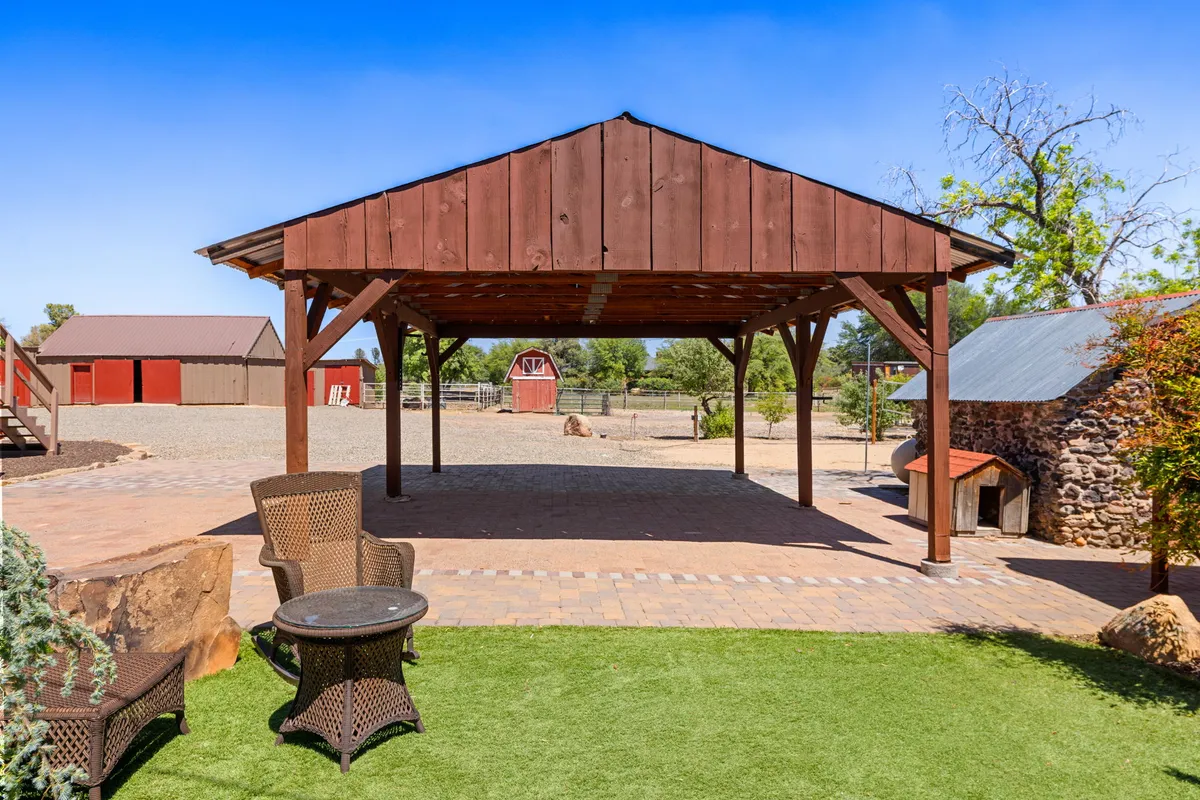 A rustic wooden pavilion surrounded by green lawns and stone accents sits near classic red barns, signaling a working ranch environment. The outdoor space invites guests to relax, host gatherings, or set out on animal adventures across the property.